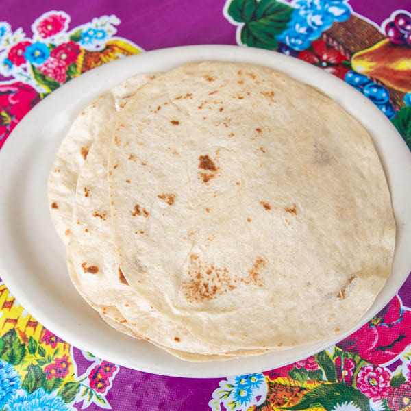 A stack of warm flour tortillas on a white plate, set against a vibrant purple floral tablecloth.