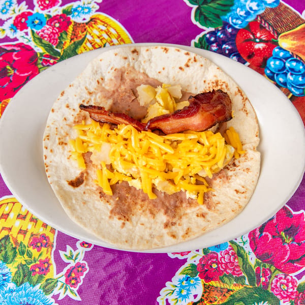 Breakfast taco with bacon, scrambled eggs, cheese, potatoes, and refried beans on a colorful tablecloth.