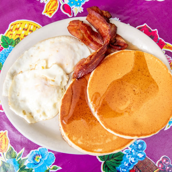 Breakfast plate with two pancakes, two fried eggs, and three strips of bacon on a colorful tablecloth.