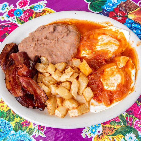A breakfast plate with bacon, refried beans, home fries, and huevos rancheros on a colorful tablecloth.