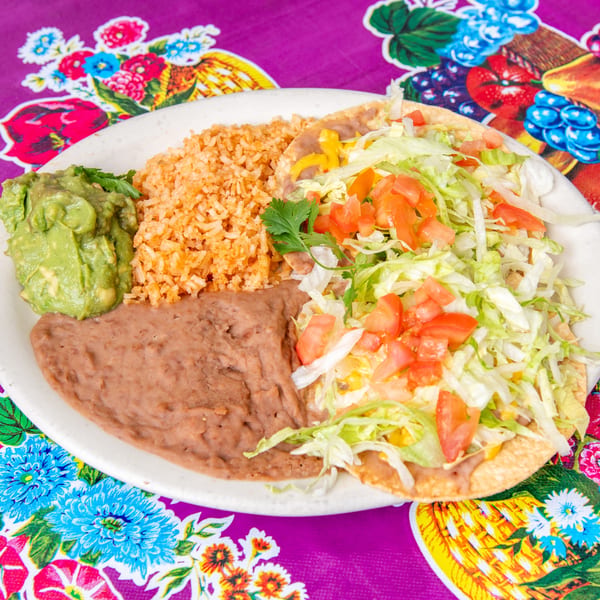 A plate of Mexican food with a tostada, rice, refried beans, and guacamole on a colorful tablecloth.