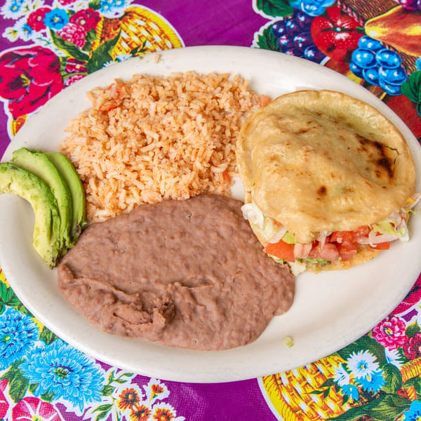 A plate of Mexican food with a gordita, rice, refried beans, and avocado slices on a floral tablecloth.