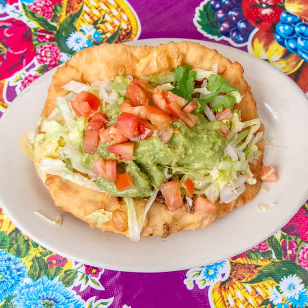 Fry bread taco with ground beef, lettuce, tomatoes, and guacamole on a colorful floral placemat.