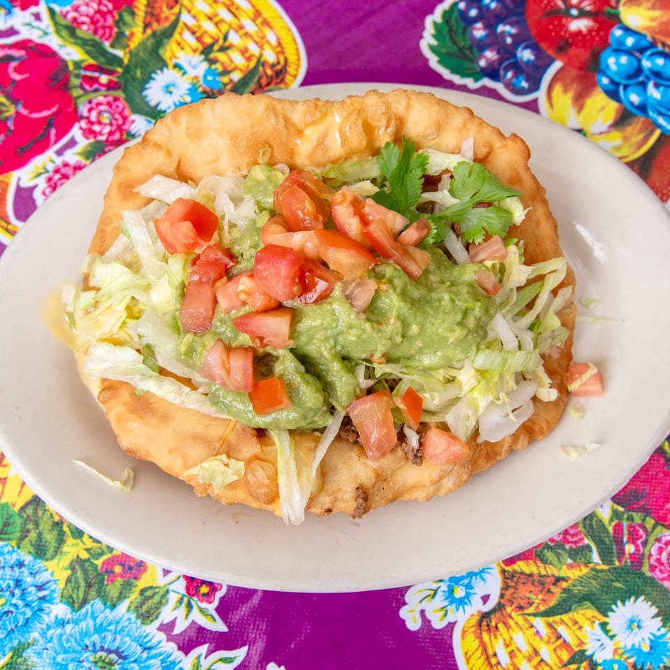 Fry bread taco with ground beef, lettuce, tomatoes, and guacamole on a colorful floral placemat.